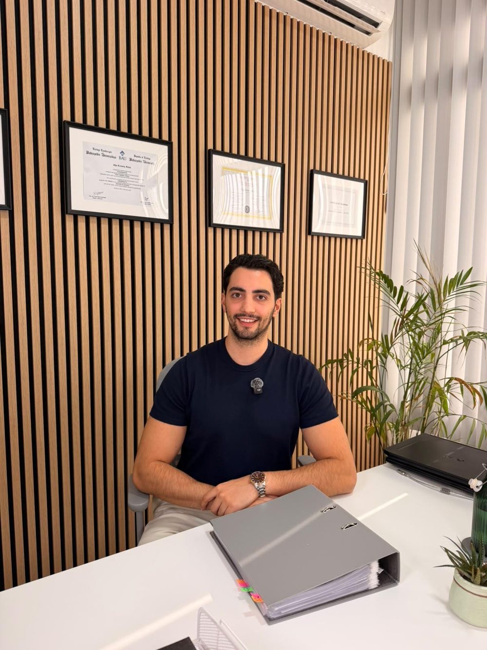 Portrait of Alp Kıvanç Riza at his desk in the clinic, with his diplomas on the wall